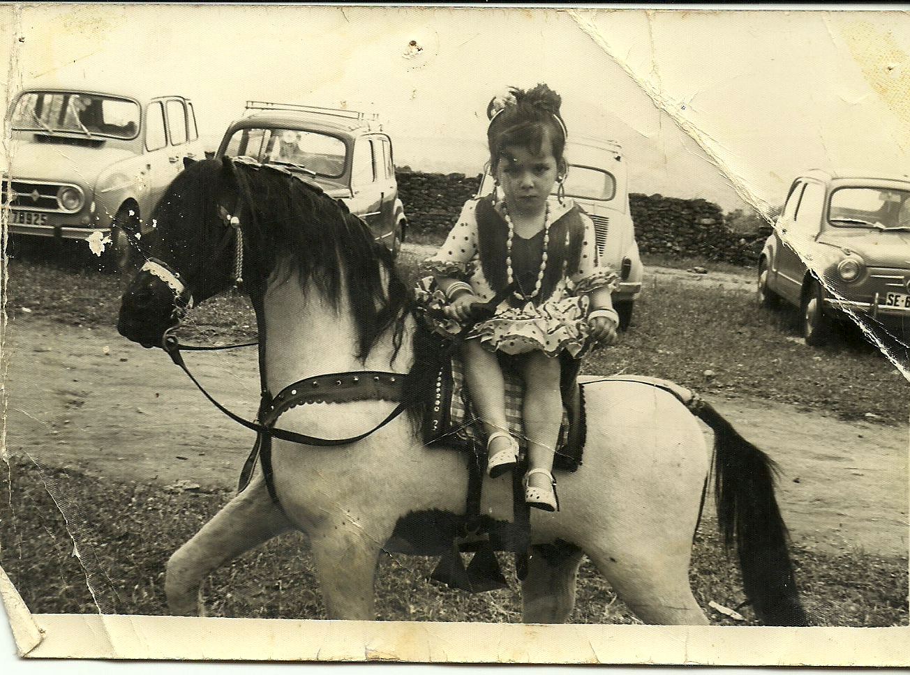 EL SEAT 600. En esta imagen vemos a Ana Carmen Andrades posando para el fotógrafo en romería en un caballo de cartón en 1969 o 1970. Detrás podemos ver el parque automovilístico de aquellos años: tres seítas y Renault "Cuatro latas". El Seat 600 fue mucho más que el primer coche de muchas familias, fue el símbolo de la época desde que comenzara a fabricarse en 1957.